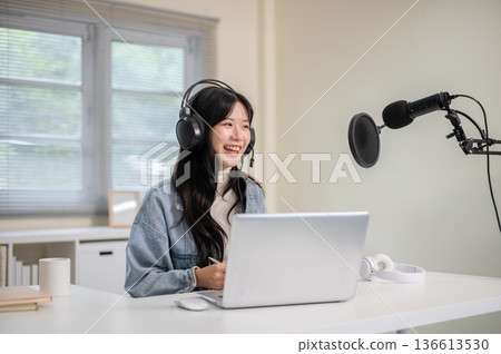 Asian woman hosting a radio show or podcast through laptop with headset and mic at home or office. Asian woman hosting a radio show or podcast through laptop with headset and mic at home or office. 136613530
