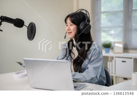 Asian woman hosting radio show or podcast through a laptop with headset and mic at home or studio. Asian woman hosting radio show or podcast through a laptop with headset and mic at home or studio. 136613548