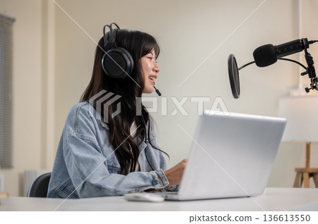 Asian woman talking hosting a radio or podcast through laptop with headset and mic at home or studio 136613550