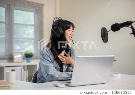 Asian woman talking hosting radio or podcast through laptop with headset and mic at home or studio. 136613553