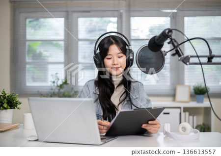 Woman reading script on clipboard as hosting radio or podcast with laptop and headset at home studio Woman reading script on clipboard as hosting radio or podcast with laptop and headset at home studio 136613557