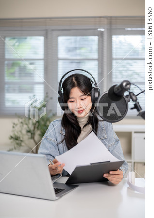 Woman reading script on clipboard while hosting radio or podcast with mic headset at home or studio. Woman reading script on clipboard while hosting radio or podcast with mic headset at home or studio. 136613560