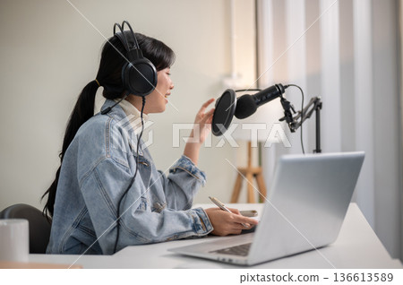 Asian woman wear headset holding mic while hosting a radio or podcast with laptop at home or studio. Asian woman wear headset holding mic while hosting a radio or podcast with laptop at home or studio. 136613589