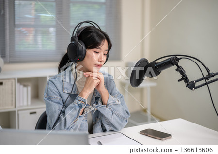 Asian woman with mic headset clasping hands looking down hosting radio or podcast at home or studio. Asian woman with mic headset clasping hands looking down hosting radio or podcast at home or studio. 136613606