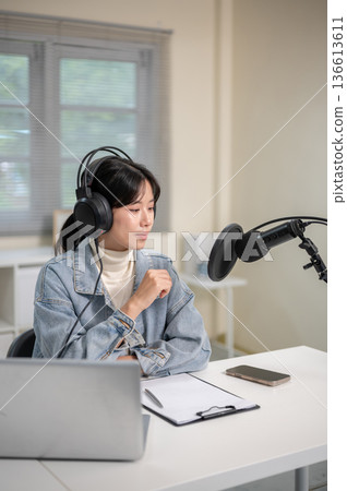 Asian woman wearing mic headset hosting a radio or podcast with laptop on table at home or studio. 136613611