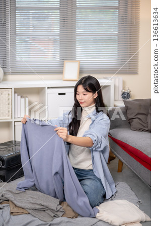 Smiling woman sitting holding cloth packing or organizing luggage for trip in bedroom or living room 136613634