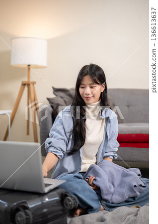 A woman holding cloth and using laptop while packing or organizing luggage in bedroom or living room 136613637