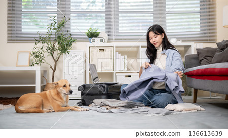 Asian woman folding cloth with laptop on luggage while packing or organizing in room with dog aside. 136613639