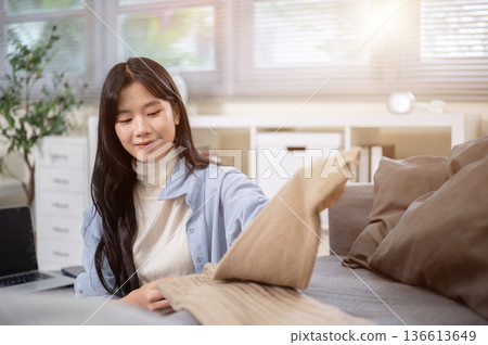 Smiling asian woman folding clothes after laundry on sofa couch in a bedroom or living room at home. 136613649
