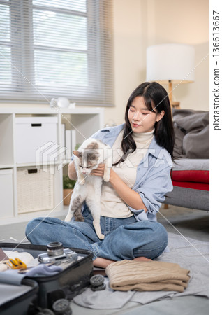 Asian woman plying with a cat while organize or packing luggage on a bedroom or living room's floor. Asian woman plying with a cat while organize or packing luggage on a bedroom or living room's floor. 136613667
