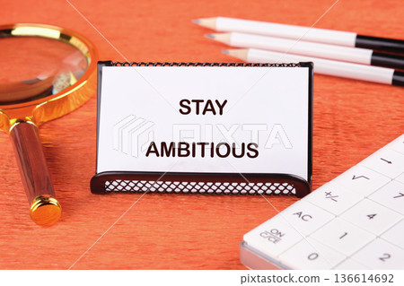 On a vibrant orange desk, a card boldly states the words Stay Ambitious, surrounded by a magnifying glass and stationery, urging focus and determination 136614692
