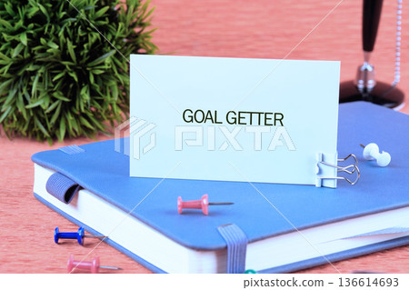 A blue notebook lies on a desk featuring a card that says Goal Getter. Colorful pins and a small plant add to the workspace atmosphere, creating inspiration 136614693