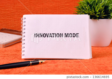 A notepad displaying the phrase innovation mode rests on a bold orange desk next to a plant, symbolizing creativity and fresh ideas ready to be explored A notepad displaying the phrase innovation mode rests on a bold orange desk next to a plant, symbolizing creativity and fresh ideas ready to be explored 136614695