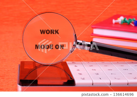 A magnifying glass displays the phrase work mode on while resting on a calculator, creating a scene of productivity against a bright orange surface A magnifying glass displays the phrase work mode on while resting on a calculator, creating a scene of productivity against a bright orange surface 136614696