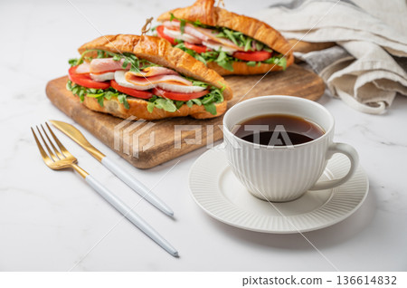 Two croissant sandwiches with ham, mozzarella cheese, tomato, arugula on a wooden board and cup of coffee 136614832