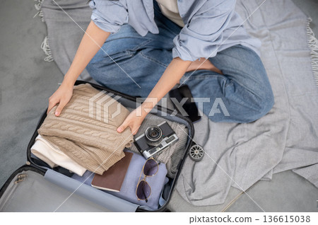 Woman putting folding cloth aside camera in luggage while organizing or packing on floor from above. Woman putting folding cloth aside camera in luggage while organizing or packing on floor from above. 136615038