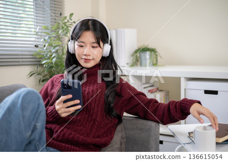 Asian woman wears headphones looking at phone sitting leaning against sofa armrest in a living room. Asian woman wears headphones looking at phone sitting leaning against sofa armrest in a living room. 136615054