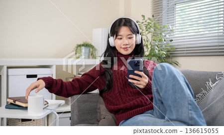 Asian woman wearings headphones holding phone sitting leaning against sofa armrest in a living room. Asian woman wearings headphones holding phone sitting leaning against sofa armrest in a living room. 136615055
