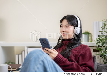 Close up of asian woman wearing headphones look away from phone while sitting on sofa in living room Close up of asian woman wearing headphones look away from phone while sitting on sofa in living room 136615058