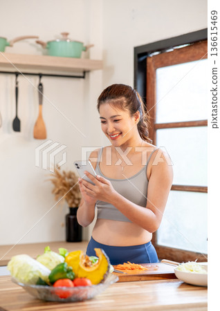 Asian woman in sportswear holding phone standing beside kitchen cooking counter with fresh vegetable 136615469