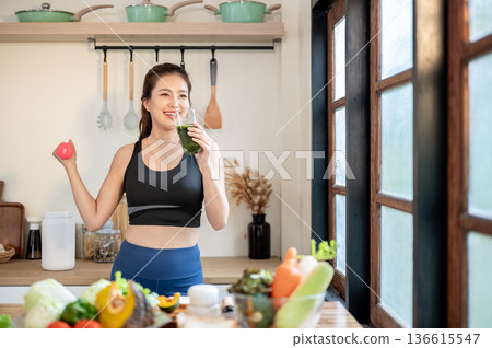 Asian sportswear woman drinking vegetable juice and doing dumbbell exercising aside kitchen counter. 136615547