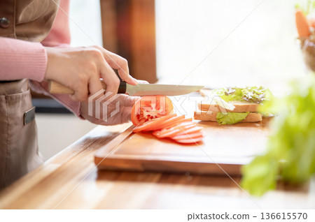 Close up of woman holding knife cutting tomato making sandwich at wooden cooking counter in kitchen. Close up of woman holding knife cutting tomato making sandwich at wooden cooking counter in kitchen. 136615570
