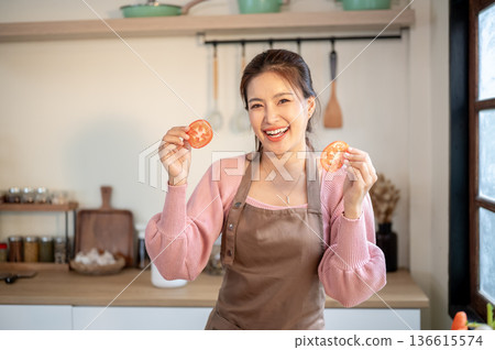 Happy asian woman wearing apron holding and showing tomato slices while cooking at kitchen counter. Happy asian woman wearing apron holding and showing tomato slices while cooking at kitchen counter. 136615574
