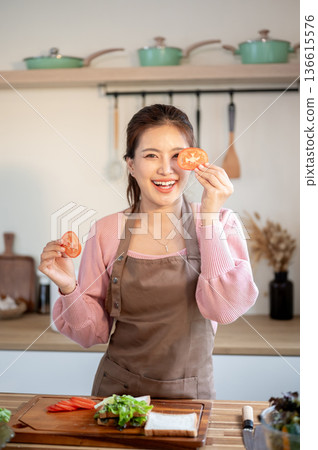 Happy asian woman wearing apron holding tomato slices making a sandwich at kitchen cooking counter. 136615576