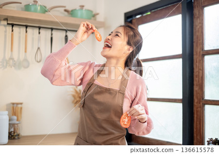 Happy asian woman wearing apron holding and eating tomato slices while cooking at kitchen counter. 136615578