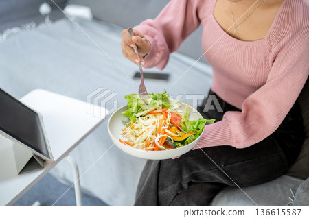 Close up of woman holding fork eating vegetable salad watching tablet sitting on sofa in living room Close up of woman holding fork eating vegetable salad watching tablet sitting on sofa in living room 136615587