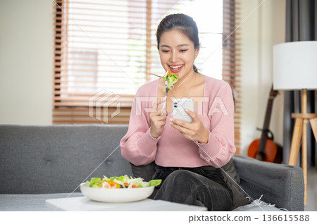 Asian woman holding fork eating vegetable salad looking at phone and sitting on sofa in living room. Asian woman holding fork eating vegetable salad looking at phone and sitting on sofa in living room. 136615588