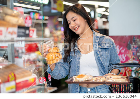 Asian woman holding shopping basket and tray while buying or looking at breads bag in supermarket. Asian woman holding shopping basket and tray while buying or looking at breads bag in supermarket. 136616044