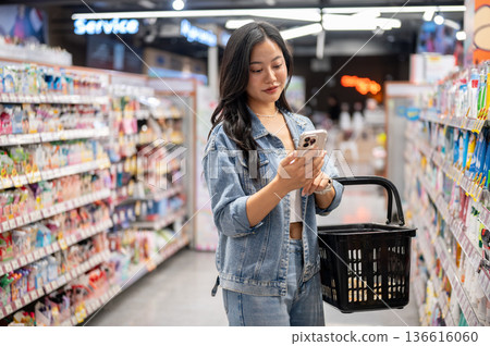 Asian woman holding a shopping basket and looking at phone while standing aside shelf in supermarket Asian woman holding a shopping basket and looking at phone while standing aside shelf in supermarket 136616060