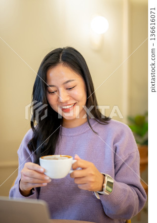Asian woman holding and looking at latte art coffee cup while sitting across laptop in cafe or shop. Asian woman holding and looking at latte art coffee cup while sitting across laptop in cafe or shop. 136616071