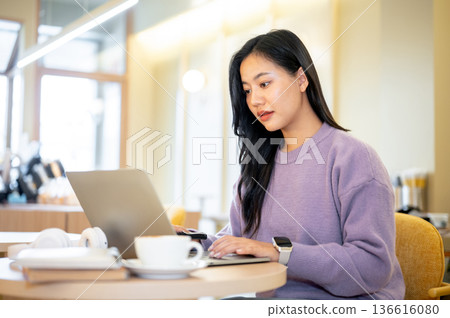 Asian woman holding pen and looking typing on laptop while sitting at table in cafe or coffee shop. 136616080