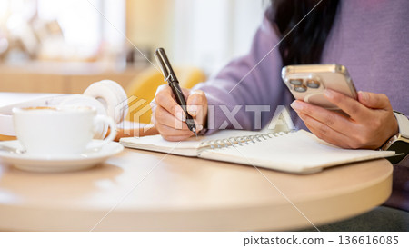 Close up of woman holding pen writing in notebook as looking at phone sitting in cafe or coffee shop 136616085