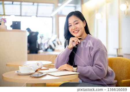 Happy asian woman holding pen under chin while sitting with notebook at table in cafe or coffee shop 136616089