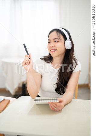 Asian woman listening to music in headphone holding pen and notebook thinking freely sits in bedroom Asian woman listening to music in headphone holding pen and notebook thinking freely sits in bedroom 136616316