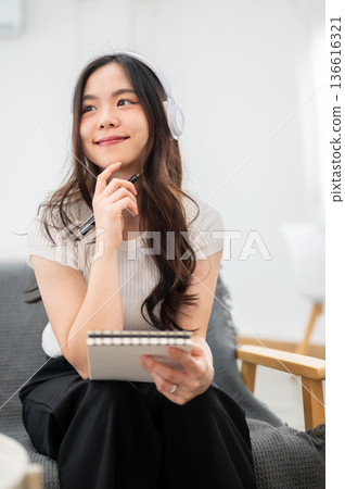 Asian woman with headphones holding notebook and pen under chin while sitting on sofa in living room Asian woman with headphones holding notebook and pen under chin while sitting on sofa in living room 136616321