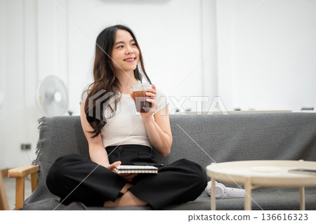 Asian woman holding notebook and drinking from a cup while sitting folded leg on sofa in living room 136616323