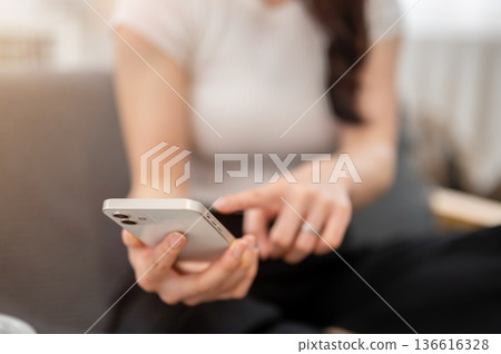 Close up of woman hand holding smartphone with finger pointing while sitting on sofa in living room Close up of woman hand holding smartphone with finger pointing while sitting on sofa in living room 136616328
