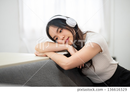 Asian woman listening to headphone music resting head on arm leaning against sofa backrest and table Asian woman listening to headphone music resting head on arm leaning against sofa backrest and table 136616329