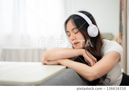 Asian woman listening to music in headphone sleeping on arms leaning against sofa backrest and table Asian woman listening to music in headphone sleeping on arms leaning against sofa backrest and table 136616330