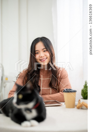 Happy asian woman looking at a black and white fur cat sitting lying on table in living room or cafe Happy asian woman looking at a black and white fur cat sitting lying on table in living room or cafe 136616369