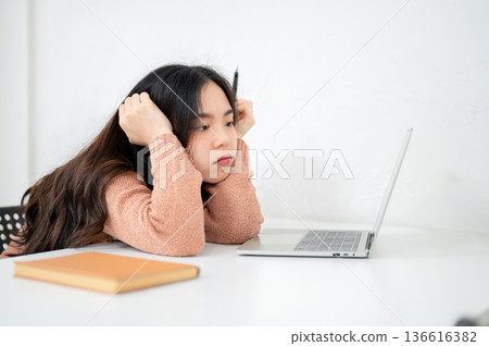 Asian woman holding head with a hand holding pen looking at laptop on table in a classroom or office 136616382
