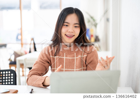 Asian woman explaining with hands and looking at a laptop on table in white room of office or cafe. Asian woman explaining with hands and looking at a laptop on table in white room of office or cafe. 136616402