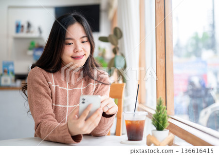 Asian woman holding and looking at phone sitting at table aside glass wall of a cafe or coffee shop. Asian woman holding and looking at phone sitting at table aside glass wall of a cafe or coffee shop. 136616415