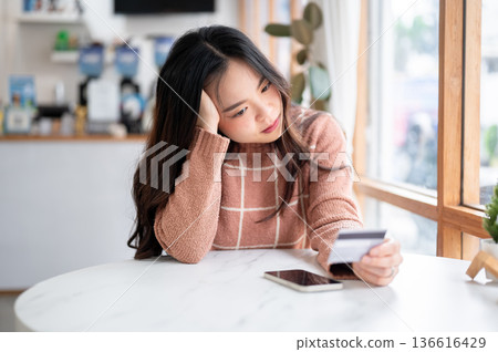 Worried asian woman holding and looking at credit card aside phone at table in a cafe or coffee shop Worried asian woman holding and looking at credit card aside phone at table in a cafe or coffee shop 136616429