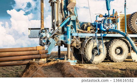 A worker operates a drilling rig at a construction site in the countryside. The rig is busy drilling into the ground as machinery and equipment are visible 136616582