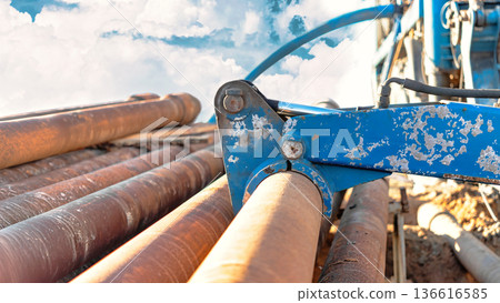 Drilling rig equipment displays metal pipes stacked on the ground. Machinery is in focus with clouds visible in the background on a clear day 136616585